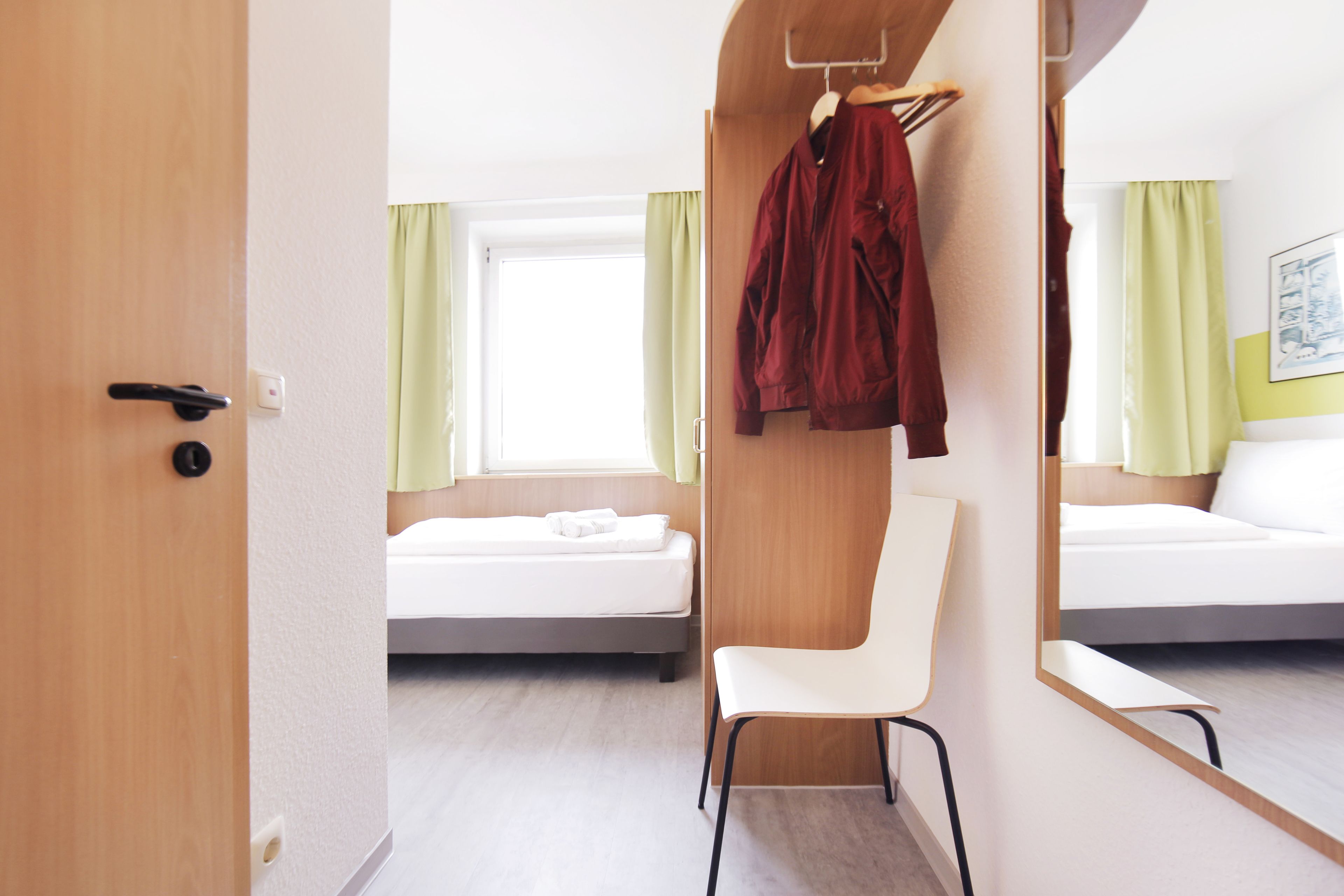 Ett hotellrum med en säng, en stol, en klädställning med en jacka på och en spegel som reflekterar rummet.