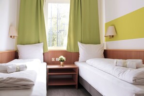 Quarto de hotel com duas camas, uma mesa de cabeceira, janela com cortinas e uma luminária na parede.