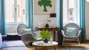 A modern living room with a sofa, two armchairs, a small table with a plant, a framed artwork of a tree, and a view of a building through a window. - ROMAC - Condotti I (Rome)