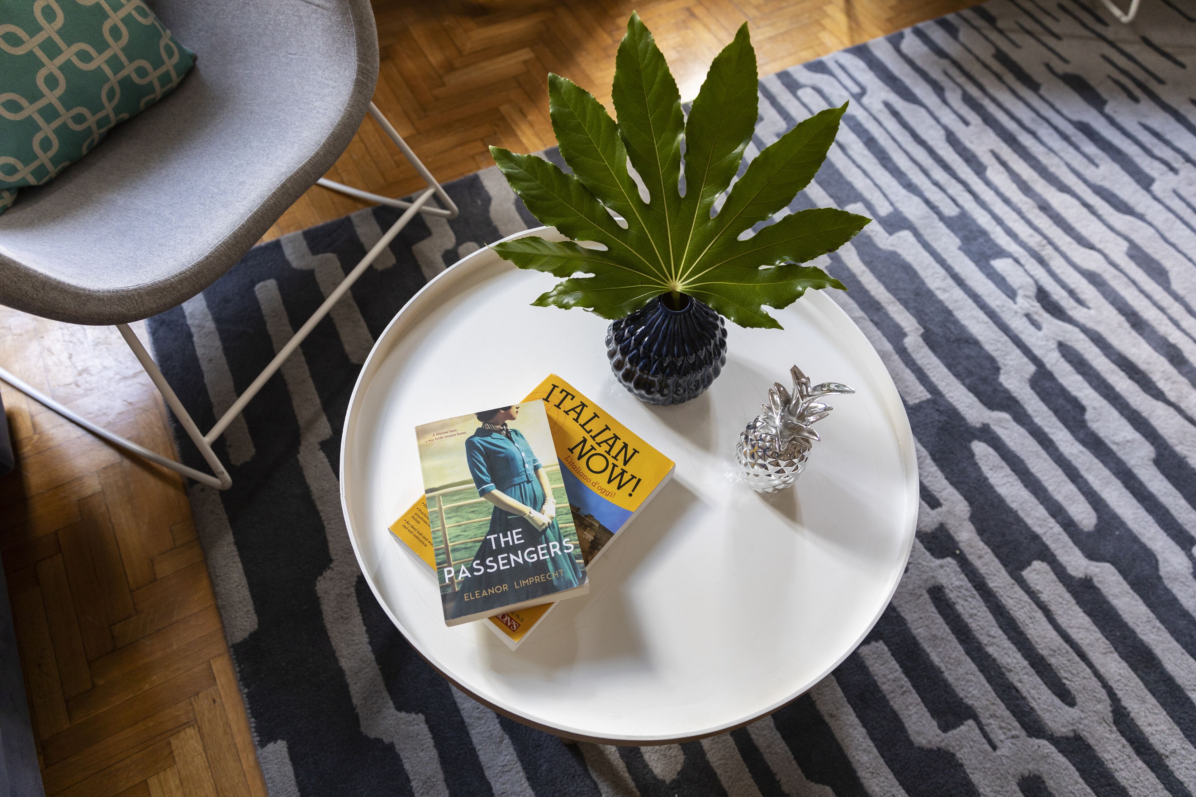 Ein weißer runder Tisch mit einer Zierpflanze, einem kleinen Blumenvasen und zwei Büchern mit den Titeln 'The Passengers' und 'Italian Now!'.