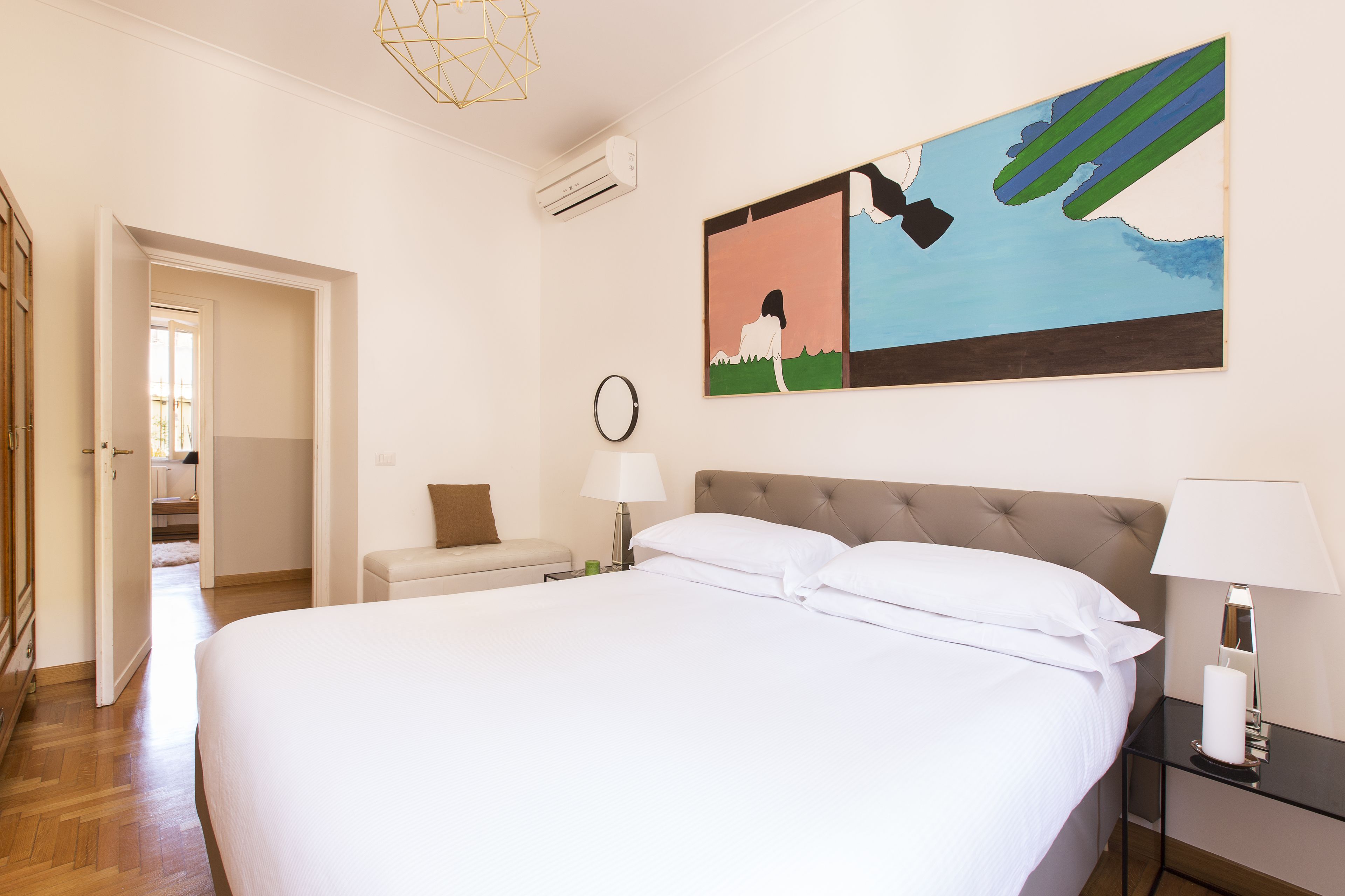 Une chambre d’hôtel avec un grand lit, une table de chevet avec une lampe, un tableau accroché au mur et une porte donnant sur une autre pièce.