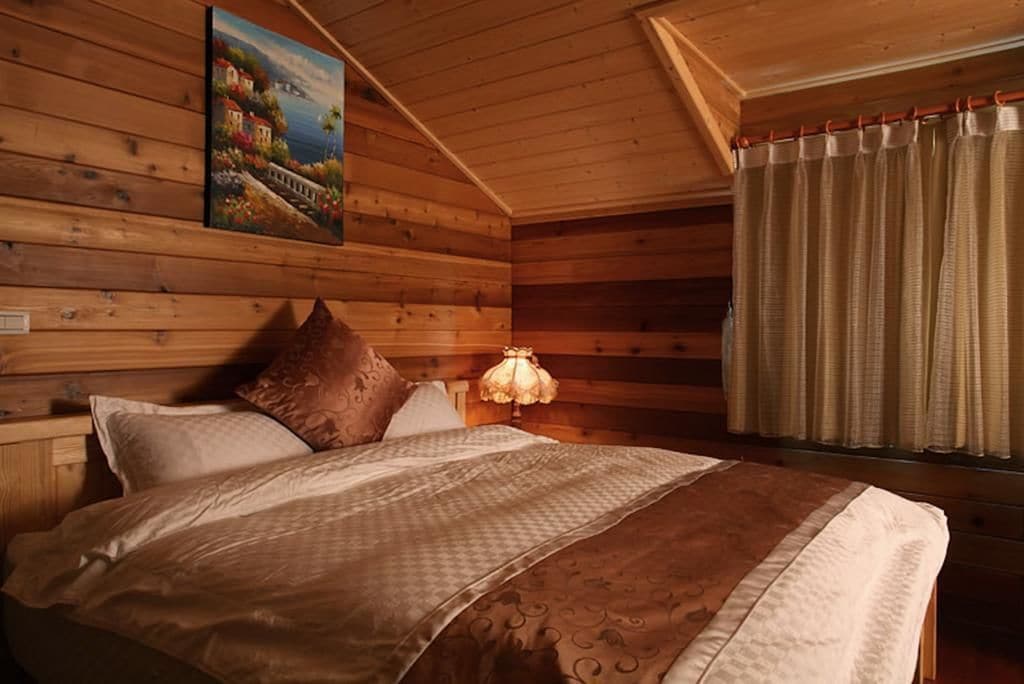 A wooden cabin bedroom with a bed, pillows, a nightstand, a lamp, and a painting on the wall.