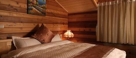 Un dormitorio de cabaña de madera con una cama, almohadas, una mesita de noche, una lámpara y un cuadro en la pared.