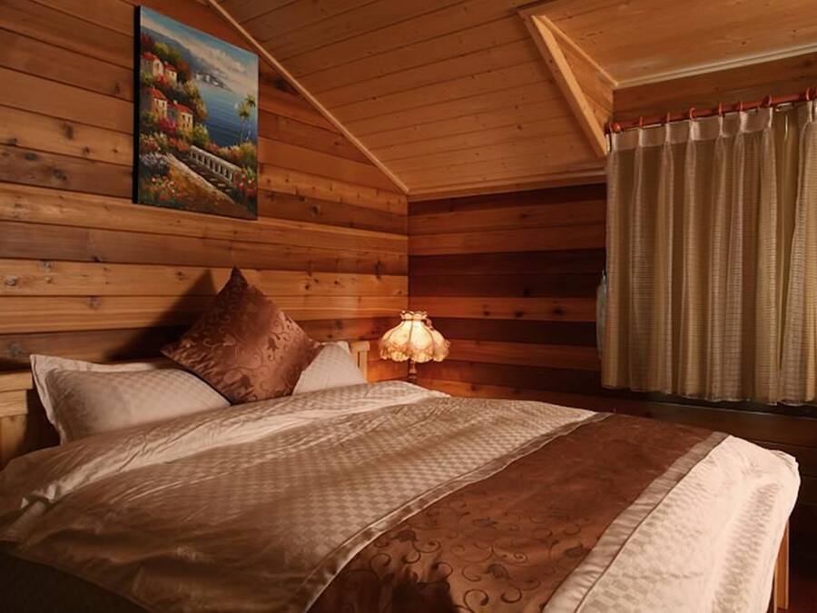 A wooden cabin bedroom with a bed, pillows, a nightstand, a lamp, and a painting on the wall.