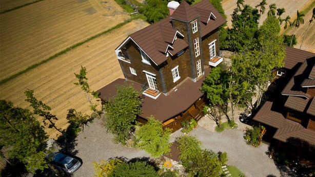 Vista aérea de uma grande casa com vários andares e uma torre imponente, cercada por um jardim bem cuidado e um campo.