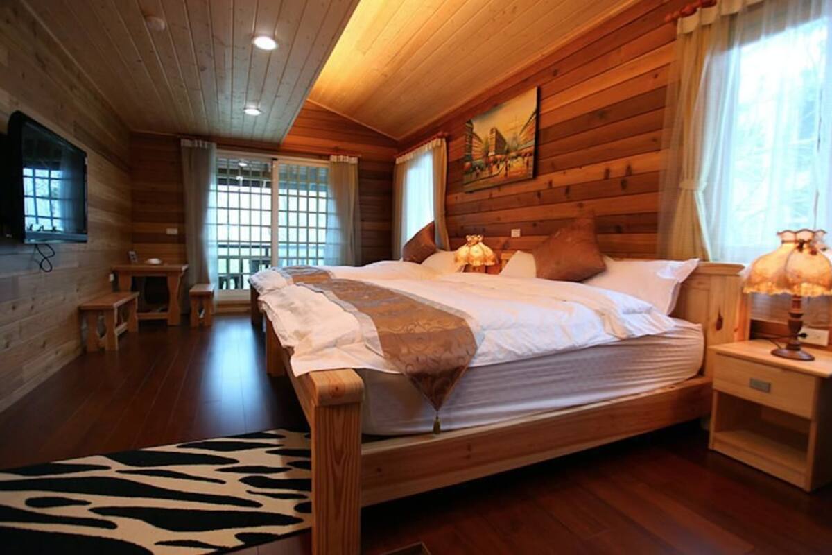 A wooden cabin room with a bed, a nightstand, a lamp, and a window with curtains.