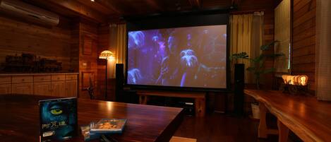 A cozy room with wooden walls and ceiling, a large screen for movie viewing, a wooden table with a DVD, a pair of glasses, and a potted plant.