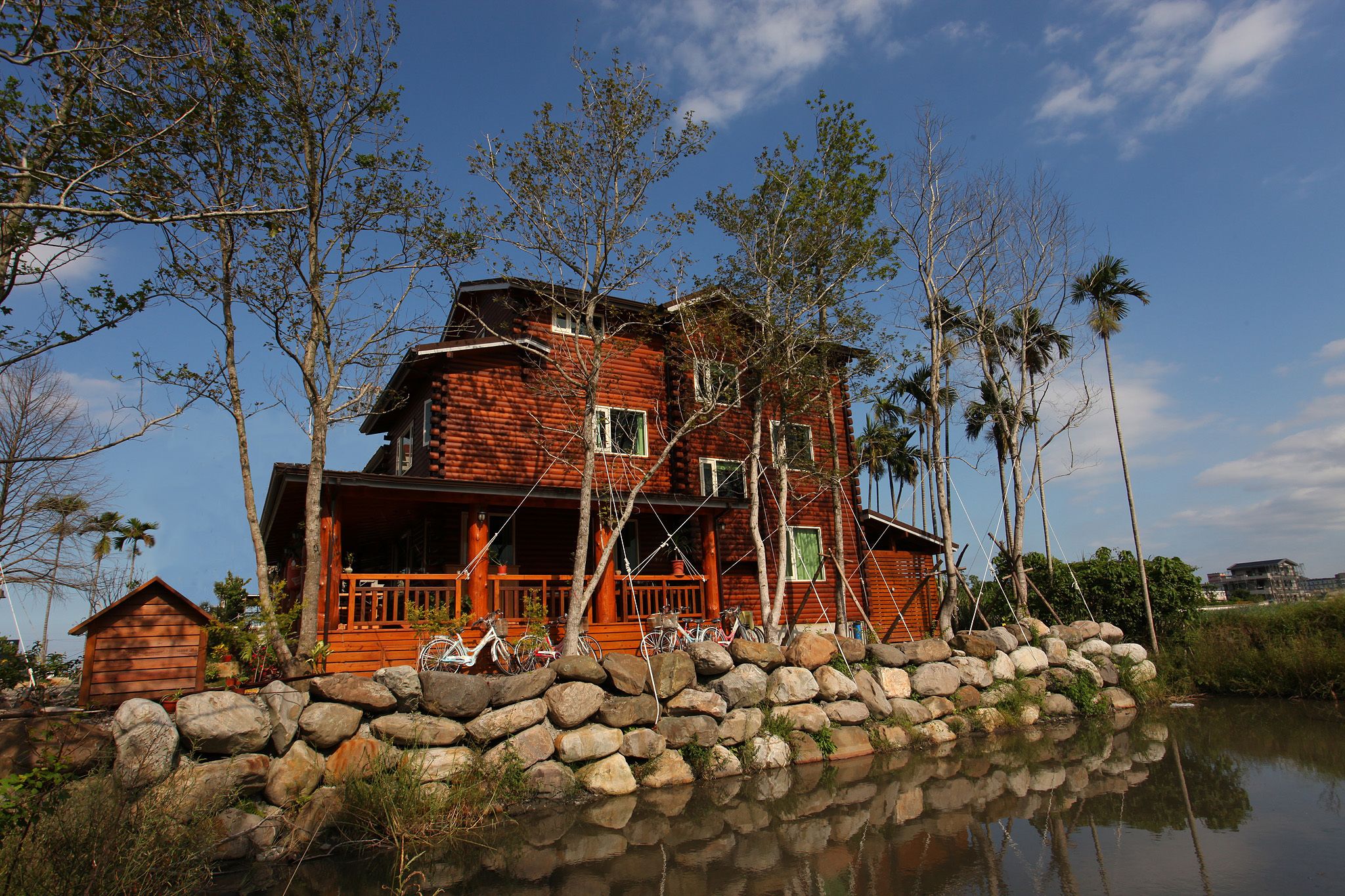 A wooden house with a porch, surrounded by rocks and trees, reflecting in a body of water.