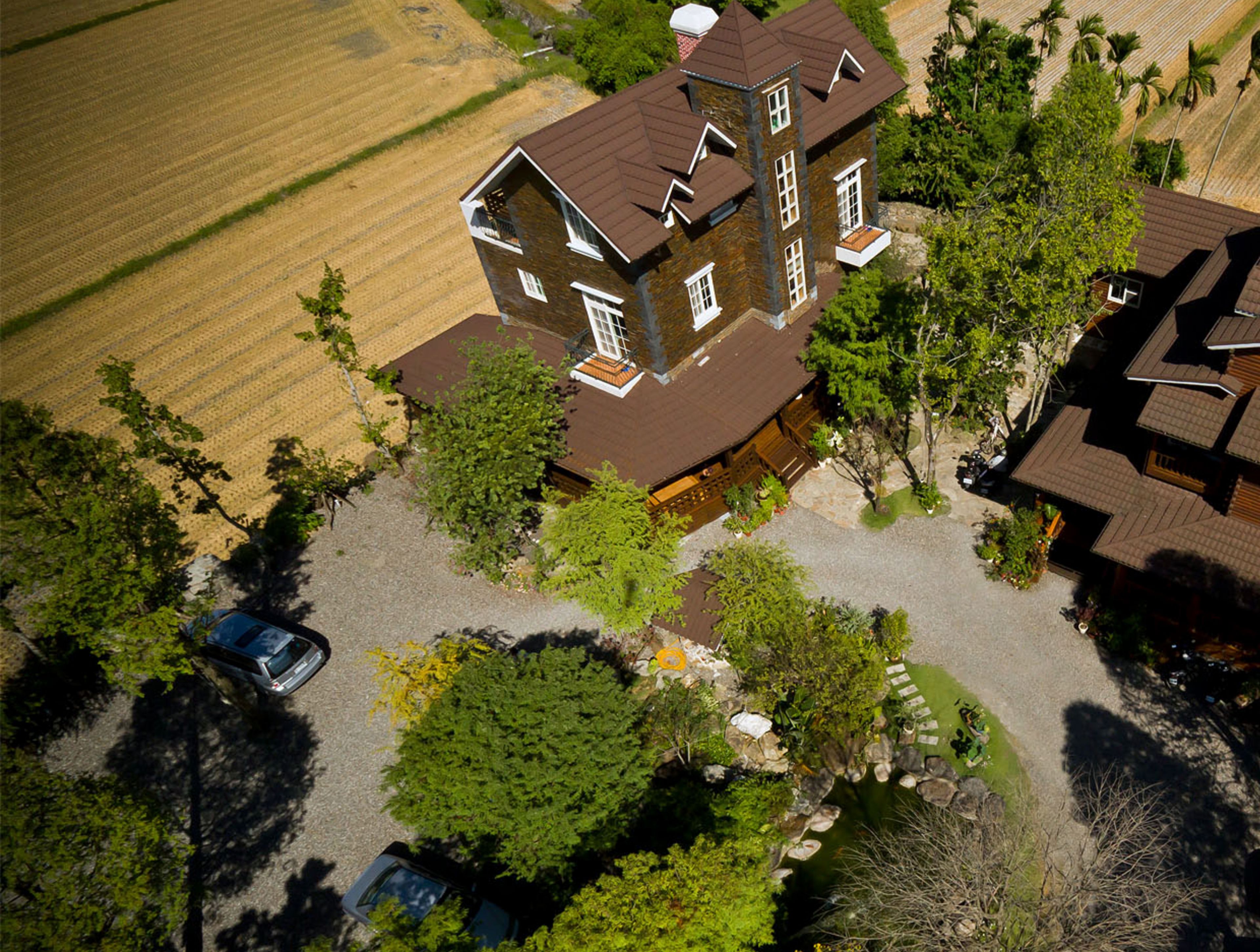 An aerial view of a large, multi-story house with a prominent tower, surrounded by a well-maintained garden and a field.