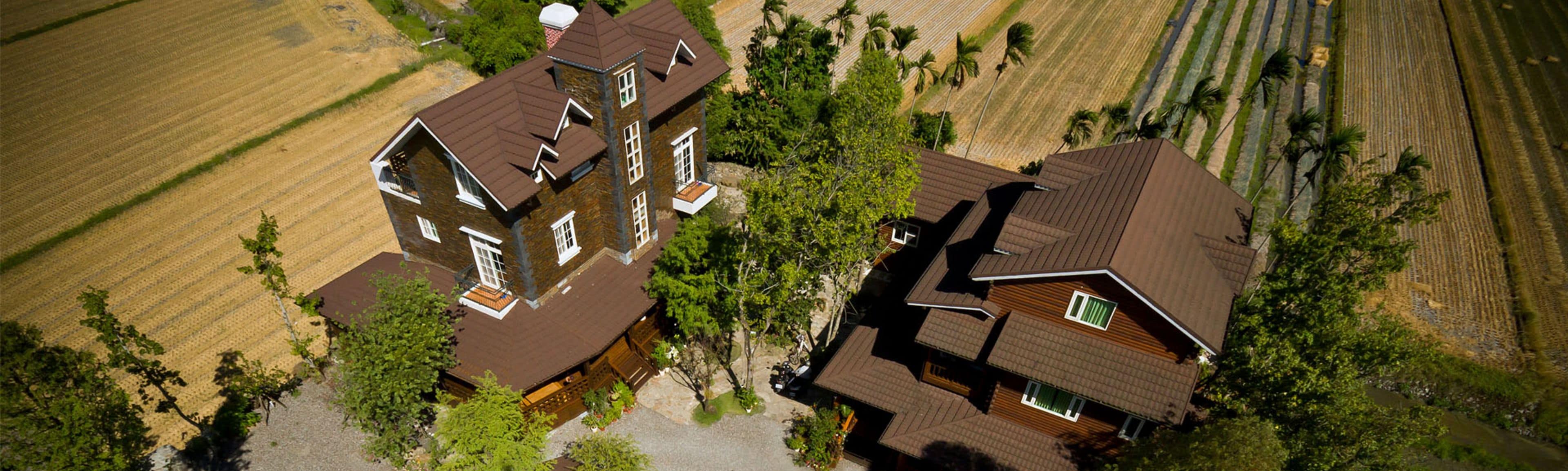 A large, multi-story house with a steep roof, surrounded by fields and trees.