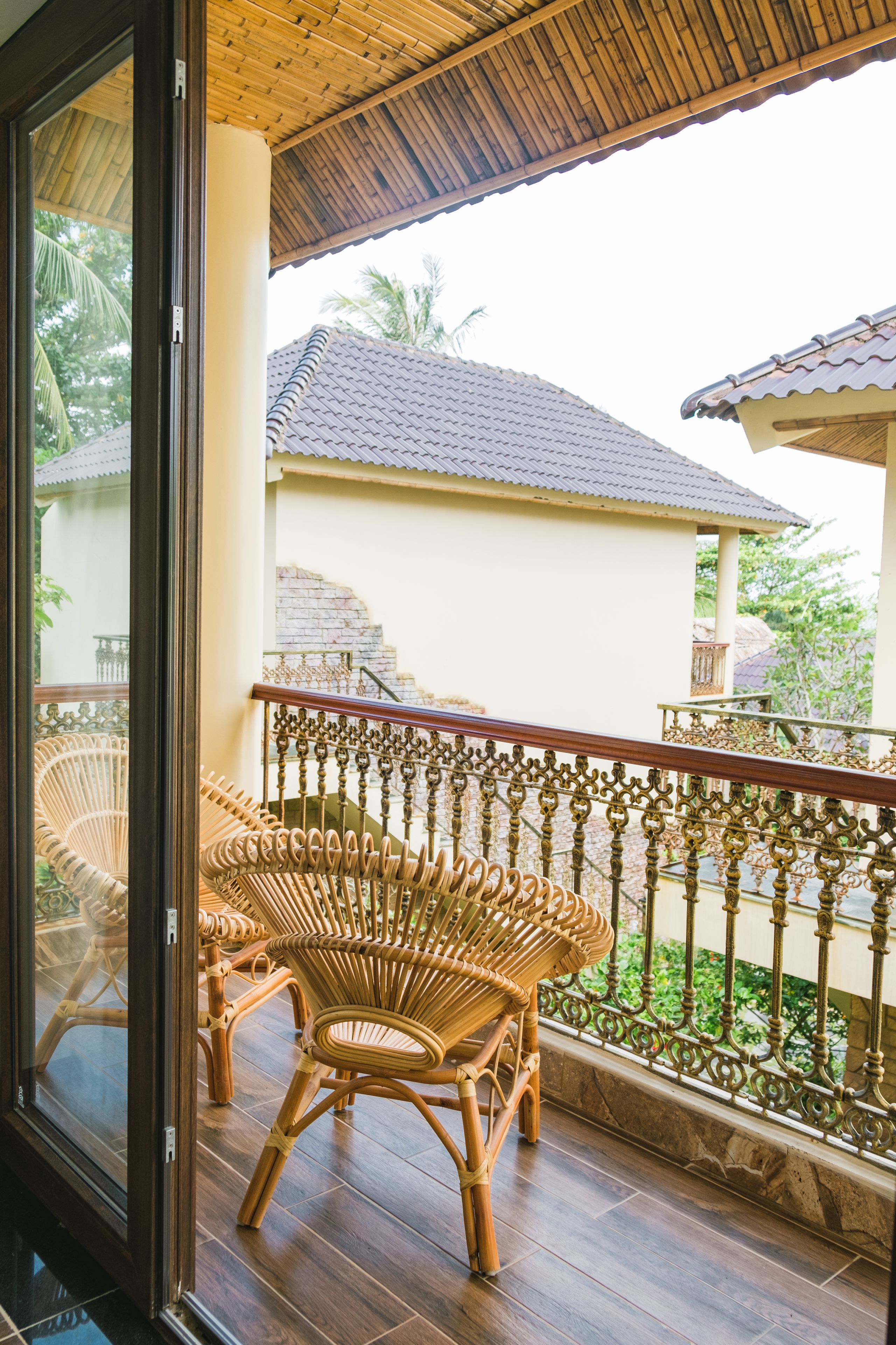 A balcony with wicker chairs, a wooden floor, and a view of a building with a tiled roof.