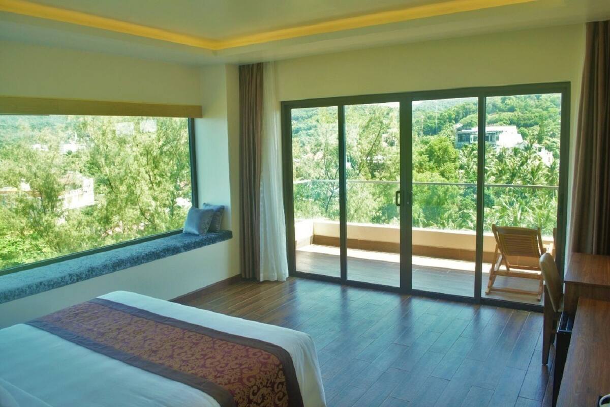 Ein Hotelzimmer mit einem großen Bett, einer Sitzbank am Fenster und einem Balkon mit Blick auf Grünflächen.