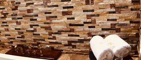 A bathroom with a stone mosaic wall, a bathtub with red petals, and towels on a black tray.