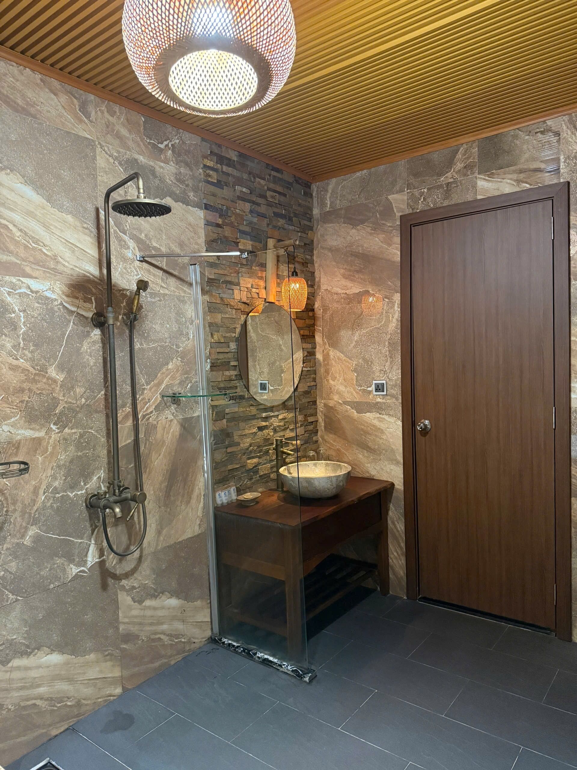 Ein Badezimmer mit Steinwand, Holztür, Duschkabine aus Glas und Waschtisch mit Becken aus Holz.