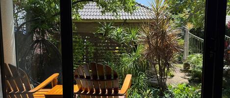 A patio area with wooden chairs and a table, surrounded by lush greenery and a tiled roof structure.