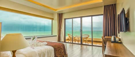 A hotel room with a bed, a desk, a chair, a TV, and a balcony with a view of the sea.