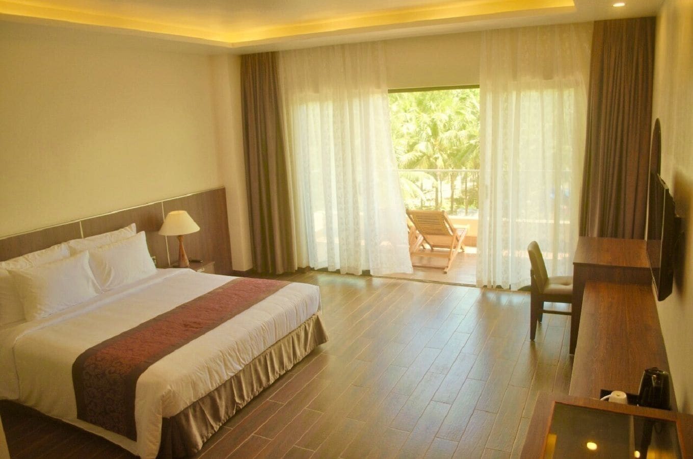 A hotel room with a large bed, a desk, a chair, and a view of the outdoors through a window with curtains.