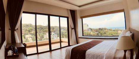 Habitación de hotel con una cama grande, una mesita de noche, un escritorio y vistas a la ciudad a través de amplios ventanales.