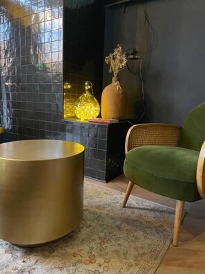 Un salon moderne avec une table ronde dorée, un fauteuil vert et des vases décoratifs sur un mur recouvert de carreaux noirs.