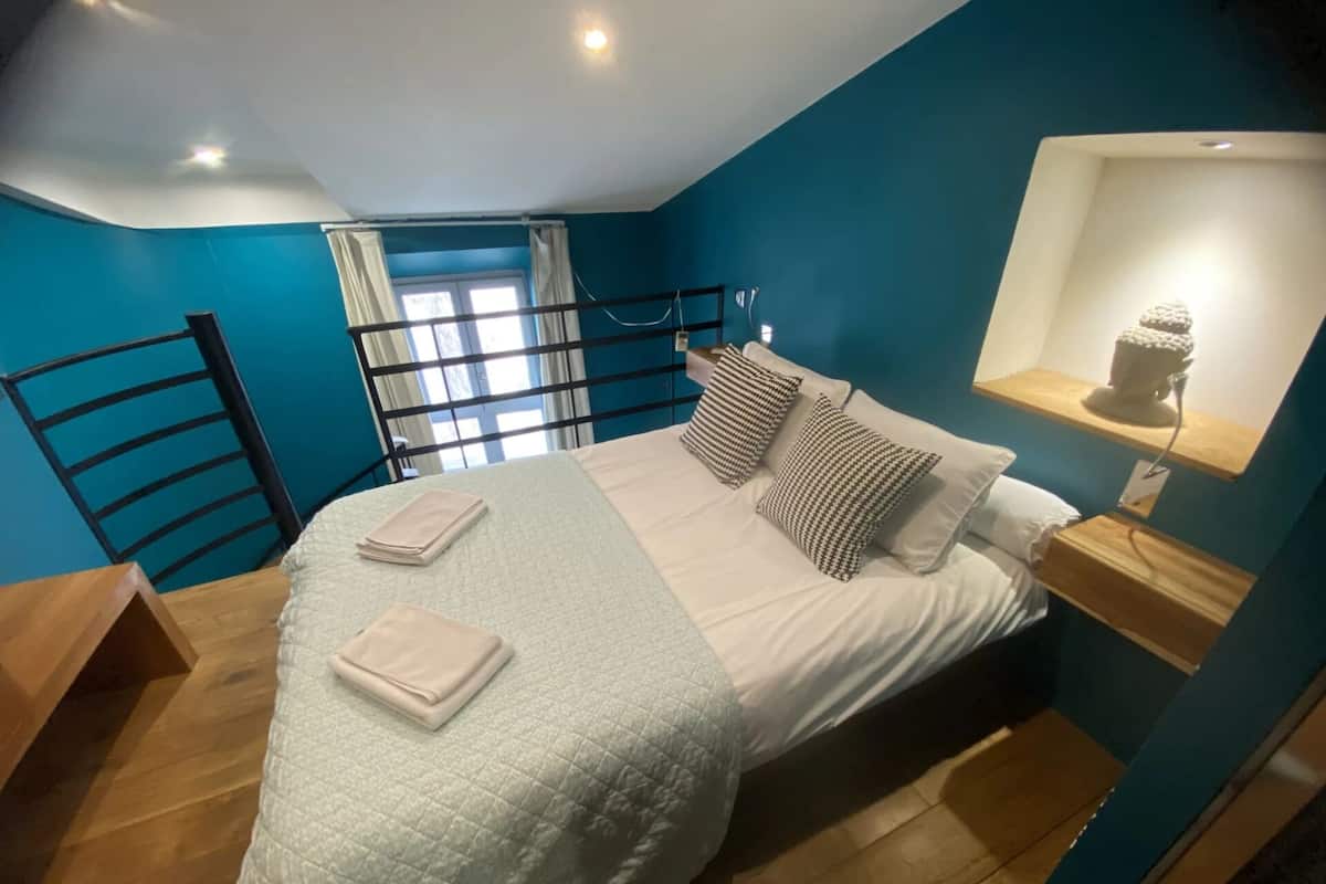Ein Schlafzimmer mit Bett, Kissen, Nachttisch, Lampe und einem Fenster mit Vorhängen.