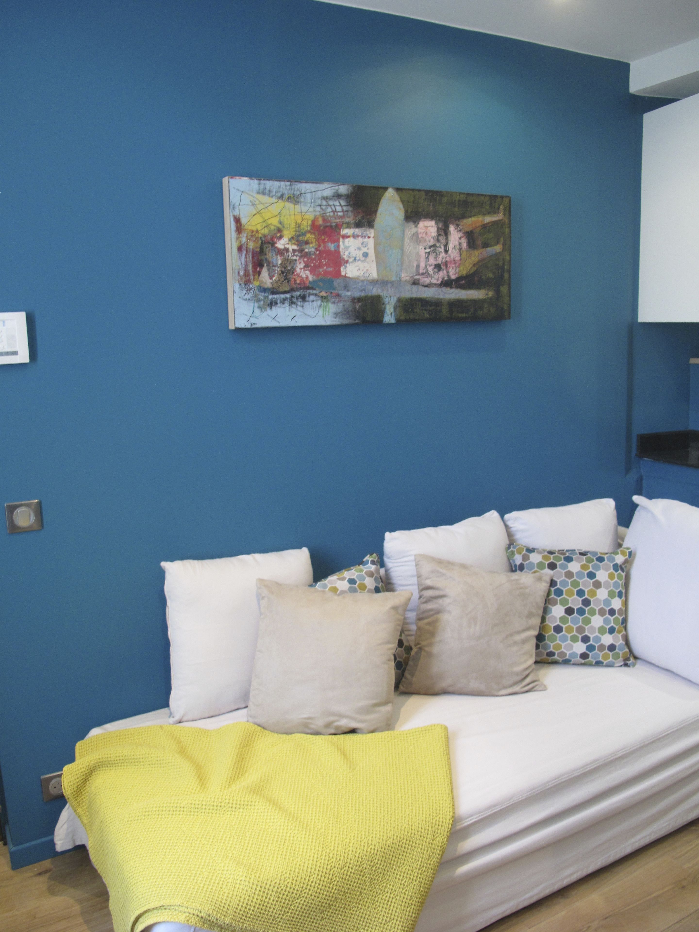 A room with a blue wall, a white sofa with multiple cushions, and a painting above the sofa.