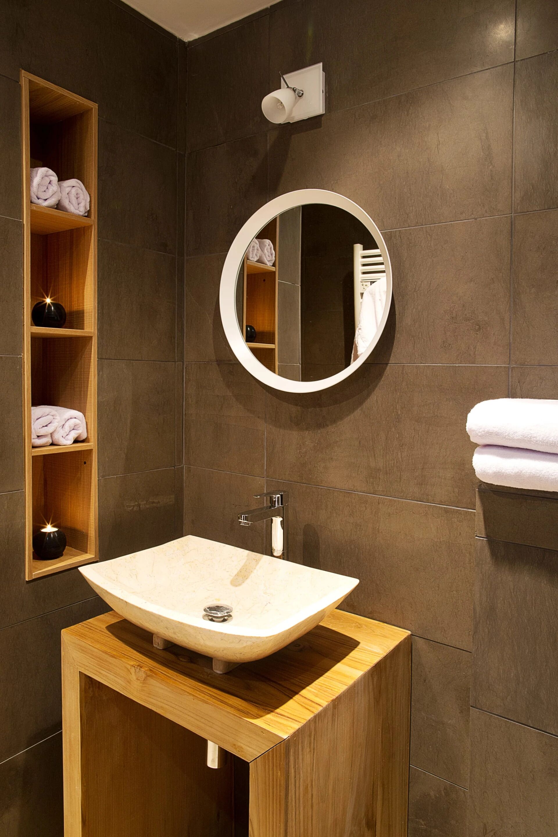Un bagno moderno con un mobile bagno in legno, uno specchio rotondo e uno scaffale integrato con asciugamani piegati.