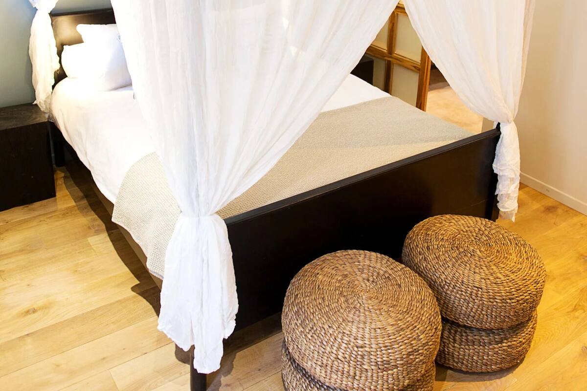 Ein Bett mit weißem Baldachin, Holzboden und zwei gewebte Hocker daneben.