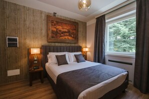 A hotel room with a large bed, wooden panel walls, a painting above the bed, two bedside lamps, a telephone, and a window with curtains. - Lake Hotel La Pieve (Pisogne)