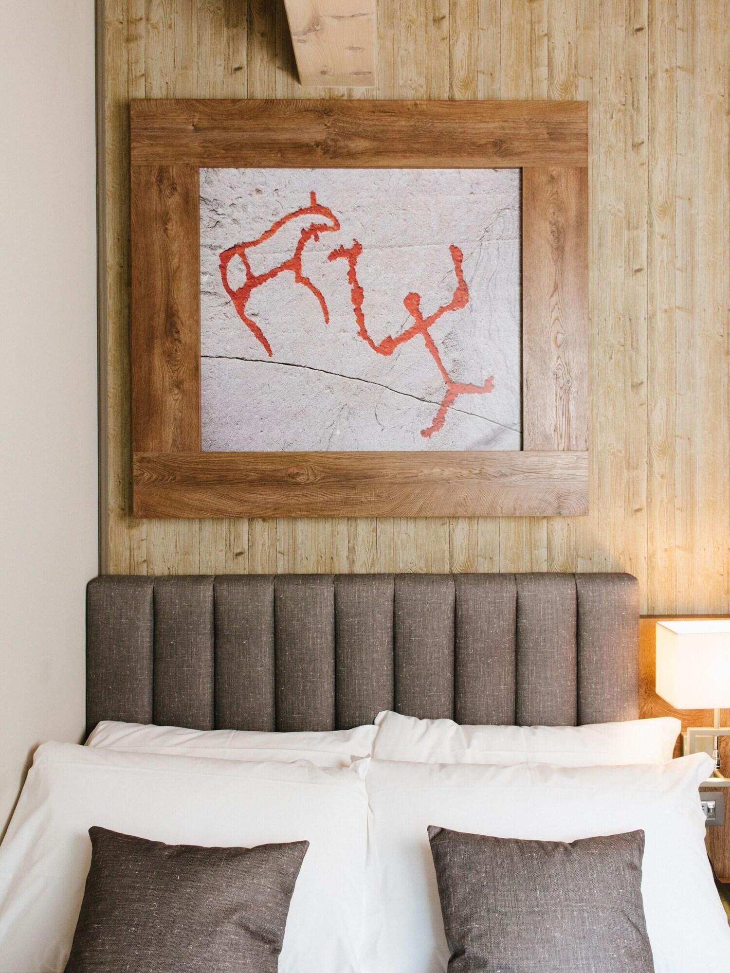 A bed with a grey headboard, white and dark grey pillows, and a framed artwork with red text on a textured wall.