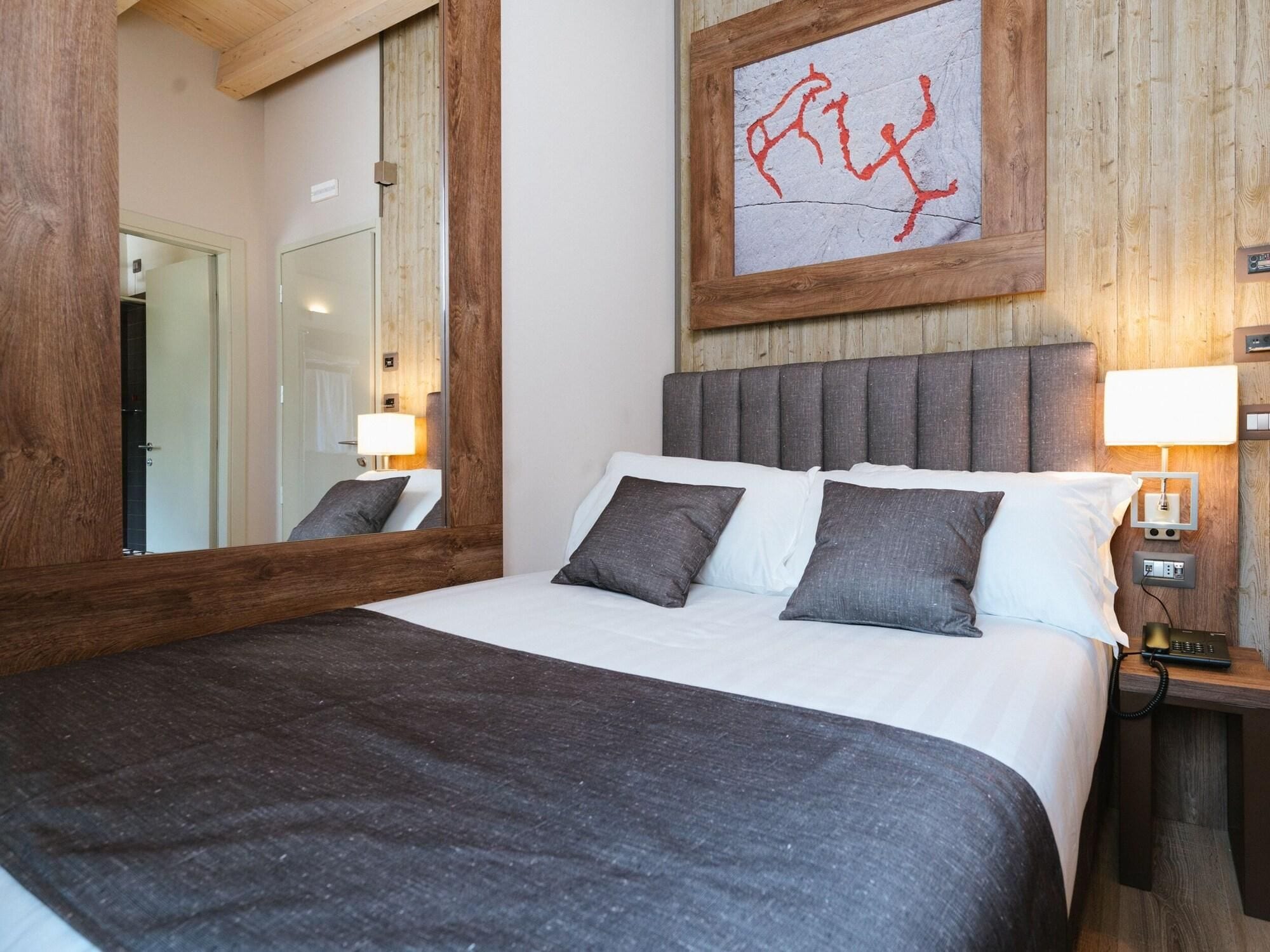 Habitación de hotel con una cama grande, cabecera de madera, un espejo, una mesita de noche con lámpara y una obra de arte colgada en la pared.
