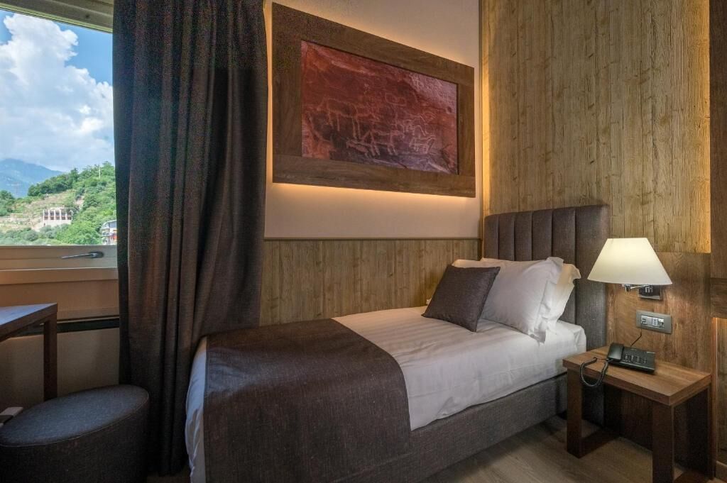 A hotel room with a bed, bedside table, lamp, and a painting on the wall.