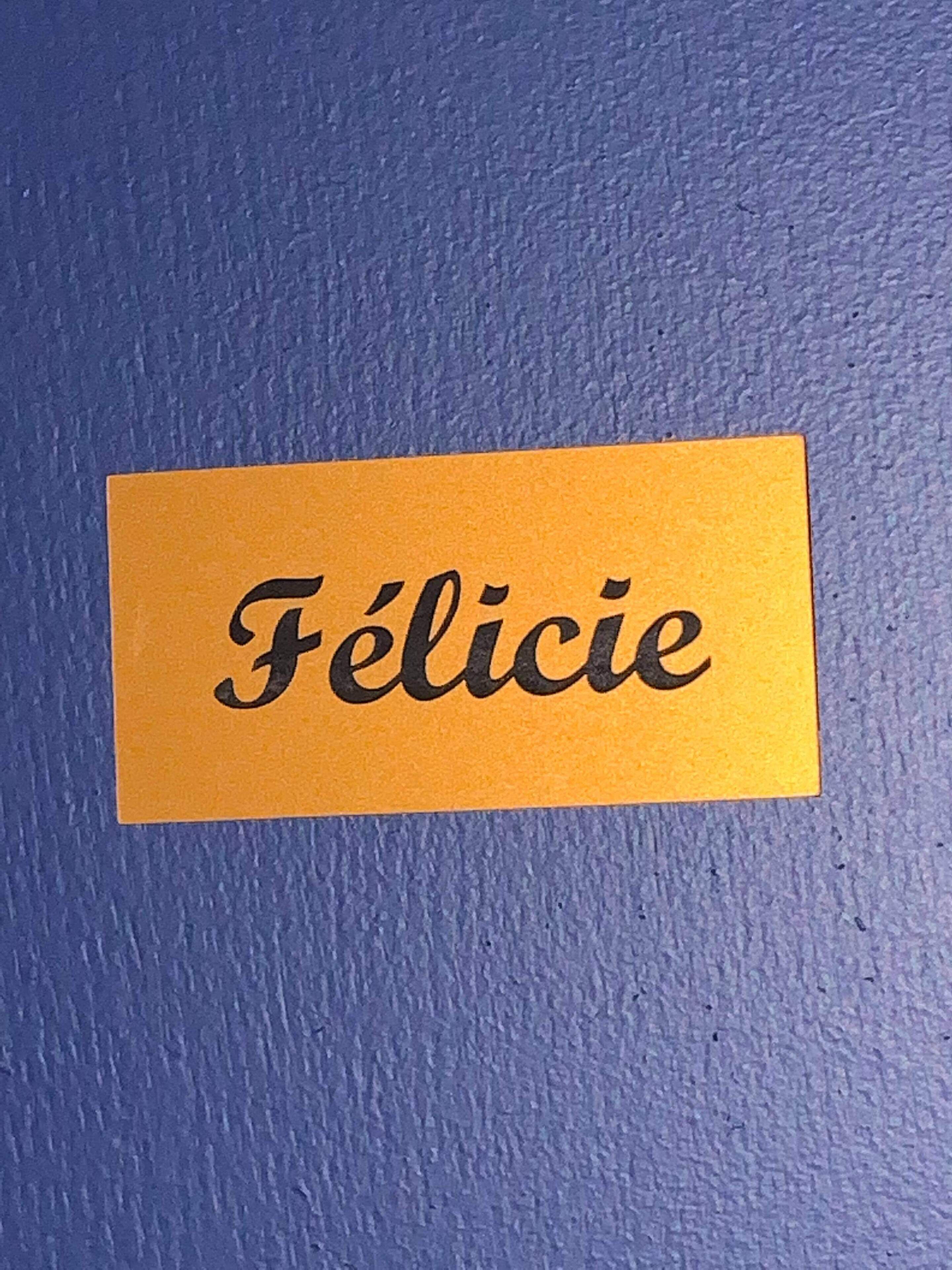 Une carte jaune sur laquelle est écrit le mot « Félicie », le tout sur un fond bleu texturé.