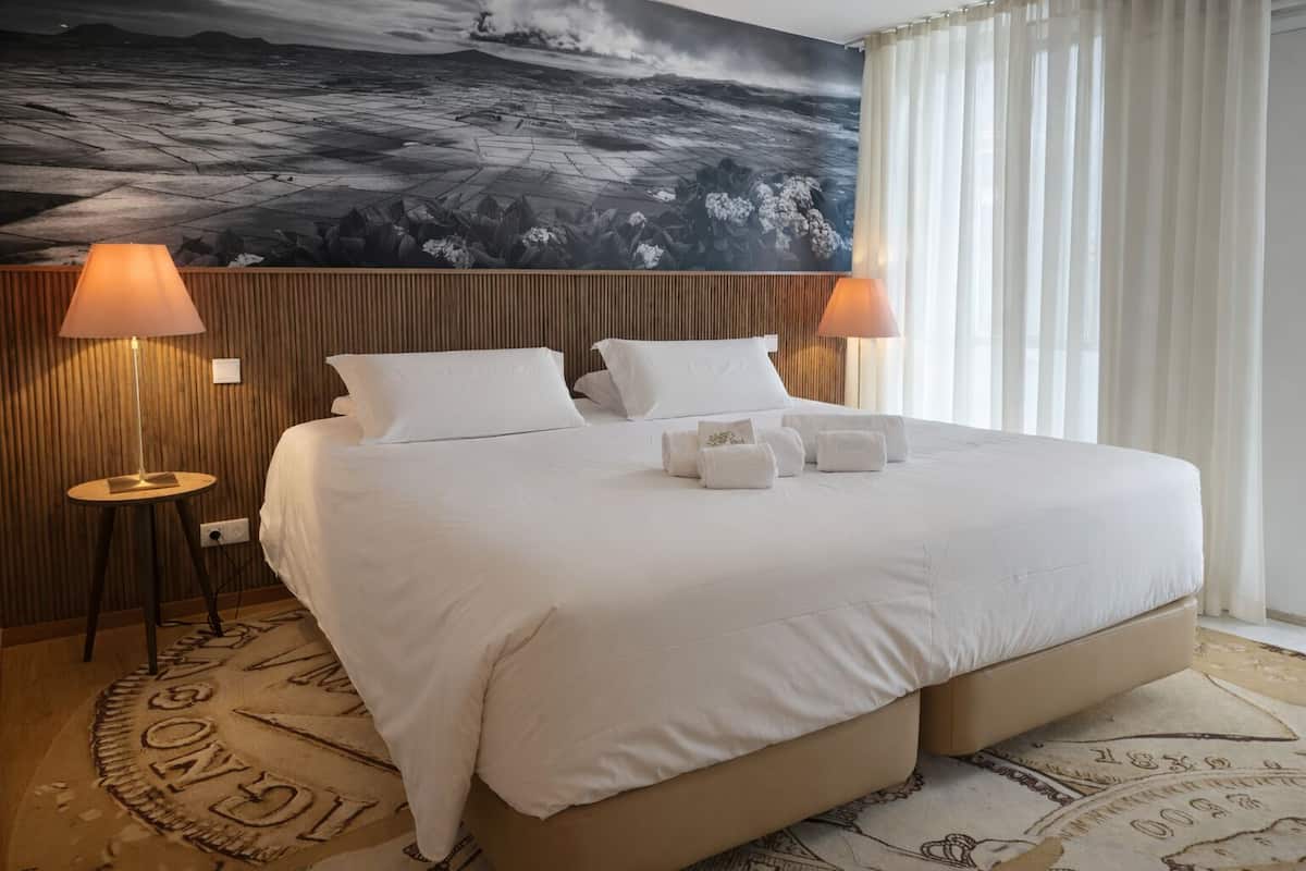 Habitación de hotel con una cama grande, dos lámparas de noche, un cuadro grande en la pared y una ventana con cortinas.