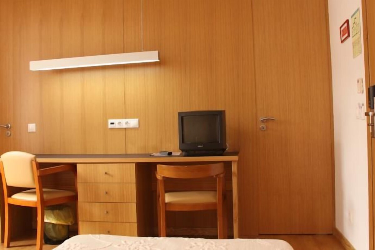 A bedroom with a wooden wardrobe, a bed, a desk with a chair, and a television.