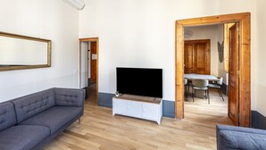 Una sala de estar con un sofá gris, un televisor sobre un mueble de madera, y un comedor con una mesa y sillas.