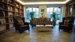 Een woonkamer met een geruite bank, twee leren fauteuils, een salontafel en een boekenkast vol ingelijste foto's en planten.