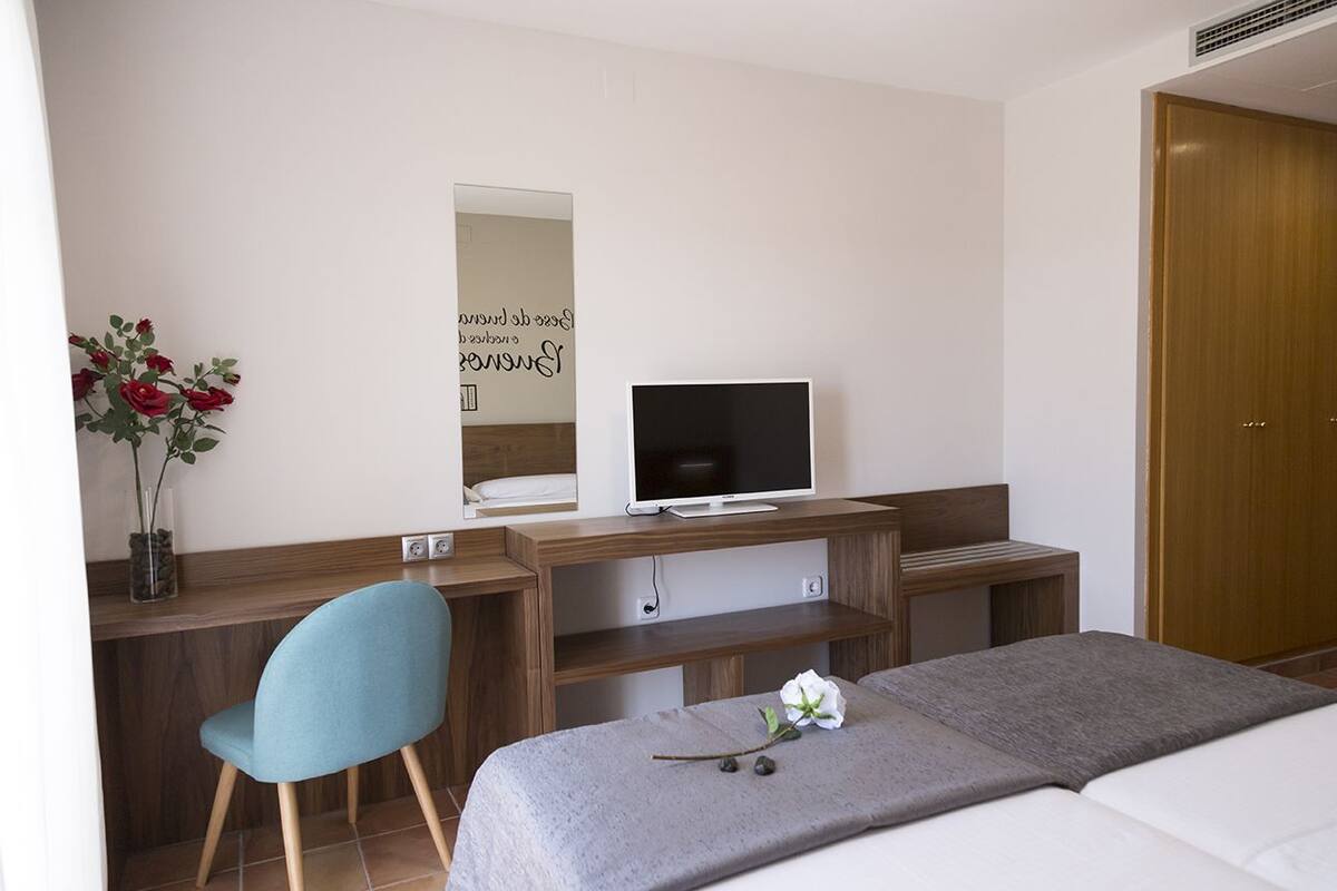 A modern hotel room with a wooden desk, a television, a bed, and a mirror on the wall.