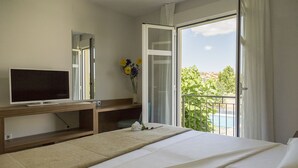 A hotel room with a bed, a TV, a desk with a computer, and a balcony with a view of greenery and a pool. - Pradaria Asador y Alojamiento (Arcas del Villar)