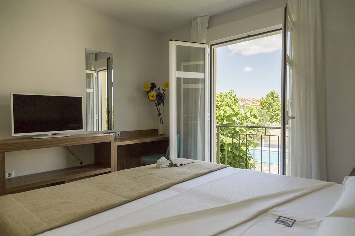 A hotel room with a bed, a TV, a desk with a computer, and a balcony with a view of greenery and a pool.