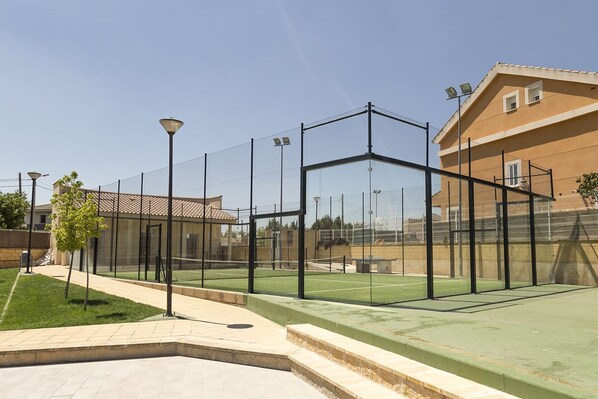 Un court de tennis avec un filet, entouré d’une clôture, et situé à côté d’un bâtiment dont le toit est recouvert de tuiles.