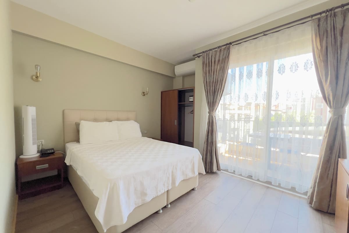 Habitación de hotel con una cama grande, una mesita de noche con lámpara, un armario y vistas al exterior a través de un amplio ventanal con cortinas.