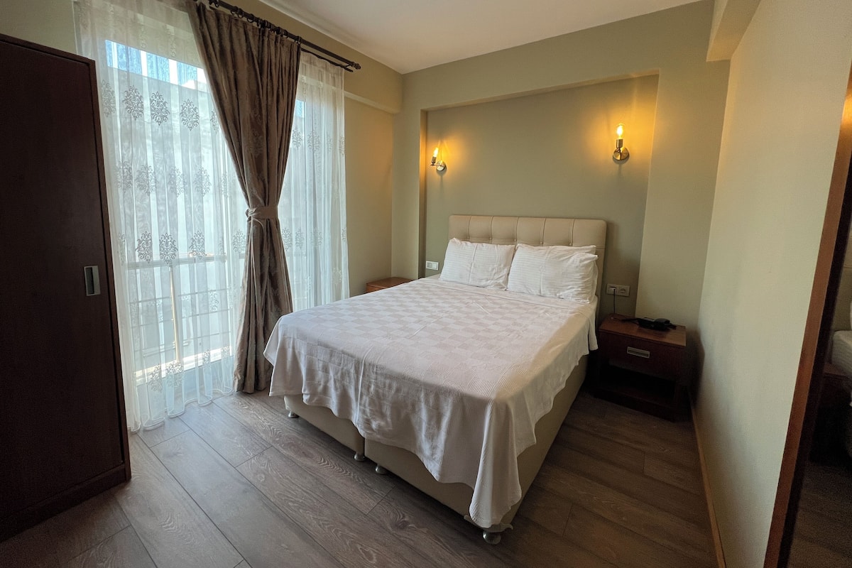 Un dormitorio con una cama grande, piso de madera, una mesita de noche y una ventana con cortinas.