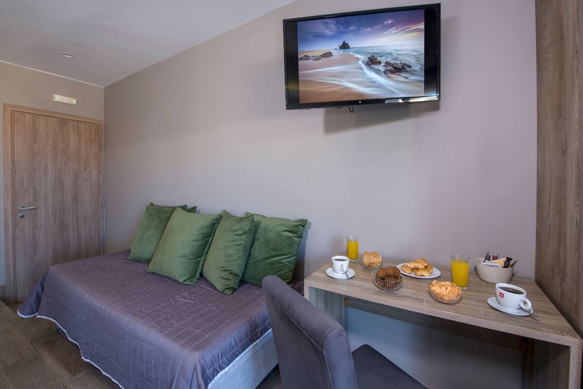 A bedroom with a bed, green pillows, a TV on the wall, a desk with breakfast items, and a wooden door.