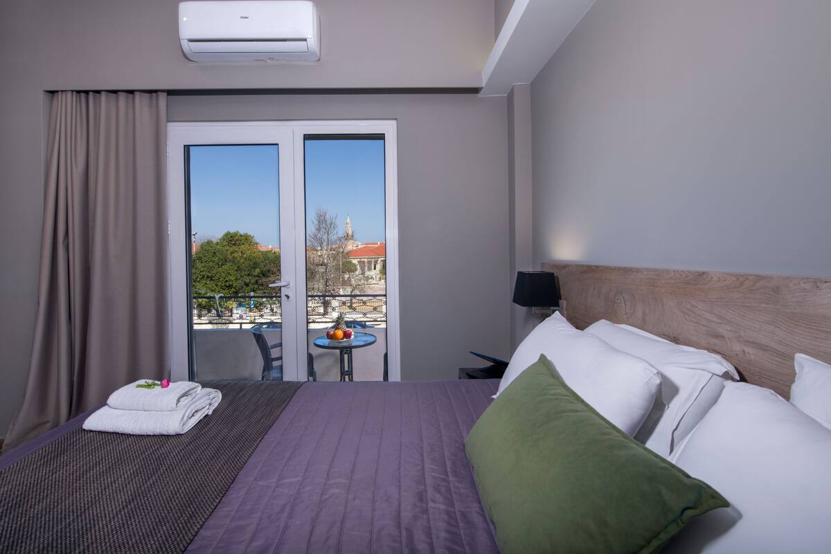 A hotel room with a bed, a small table with fruits, a balcony with a view, and an air conditioning unit.