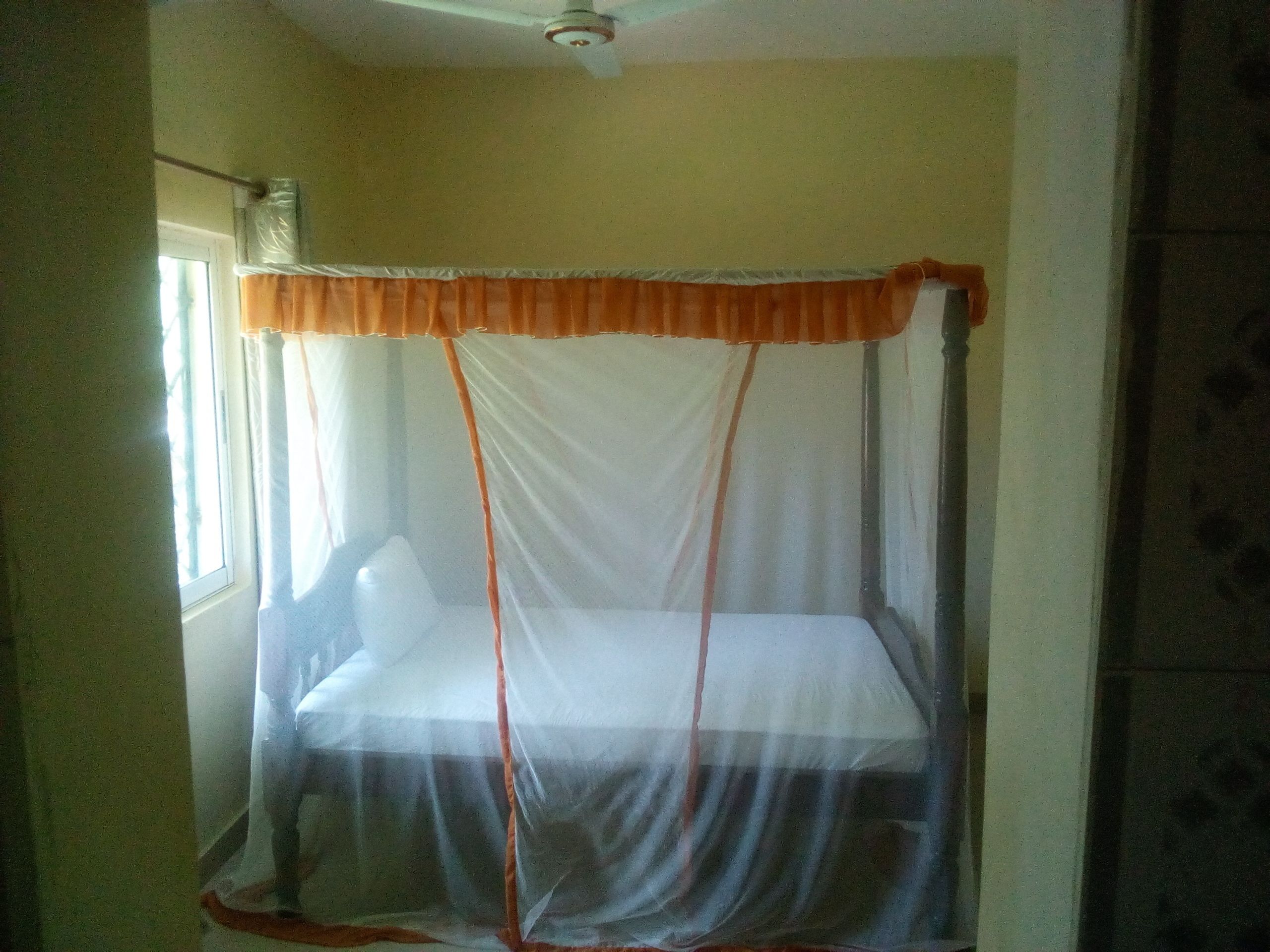 Une chambre avec un lit recouvert d’une moustiquaire, une fenêtre avec des rideaux et un ventilateur au plafond.
