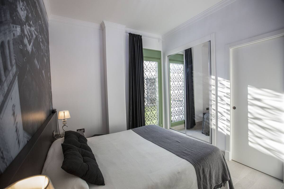 Una habitación de hotel moderna con una cama bien hecha, una lámpara de noche y un amplio ventanal con cortinas.