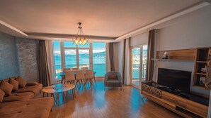 Una sala de estar moderna con un televisor grande, piso de madera, un sofá, una mesa de comedor con sillas y vista al mar.