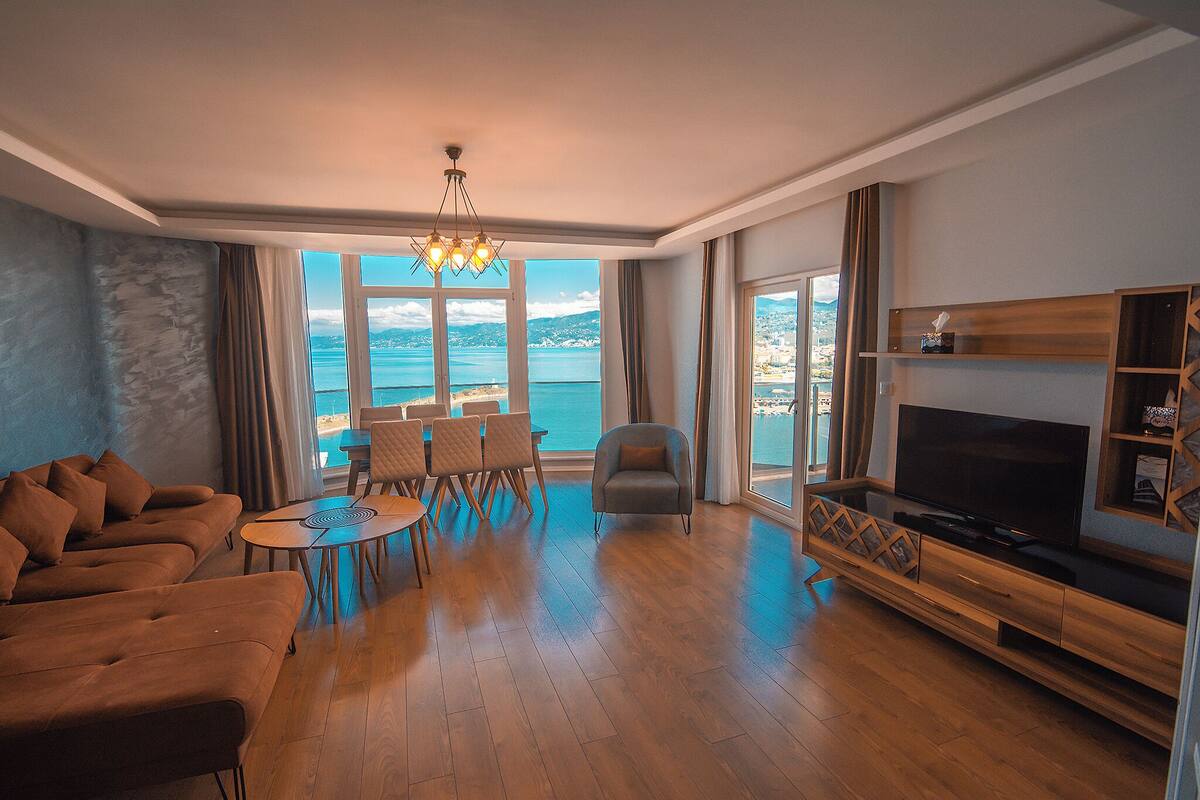 A modern living room with a large TV, wooden flooring, a sofa, a dining table with chairs, and a view of the sea.