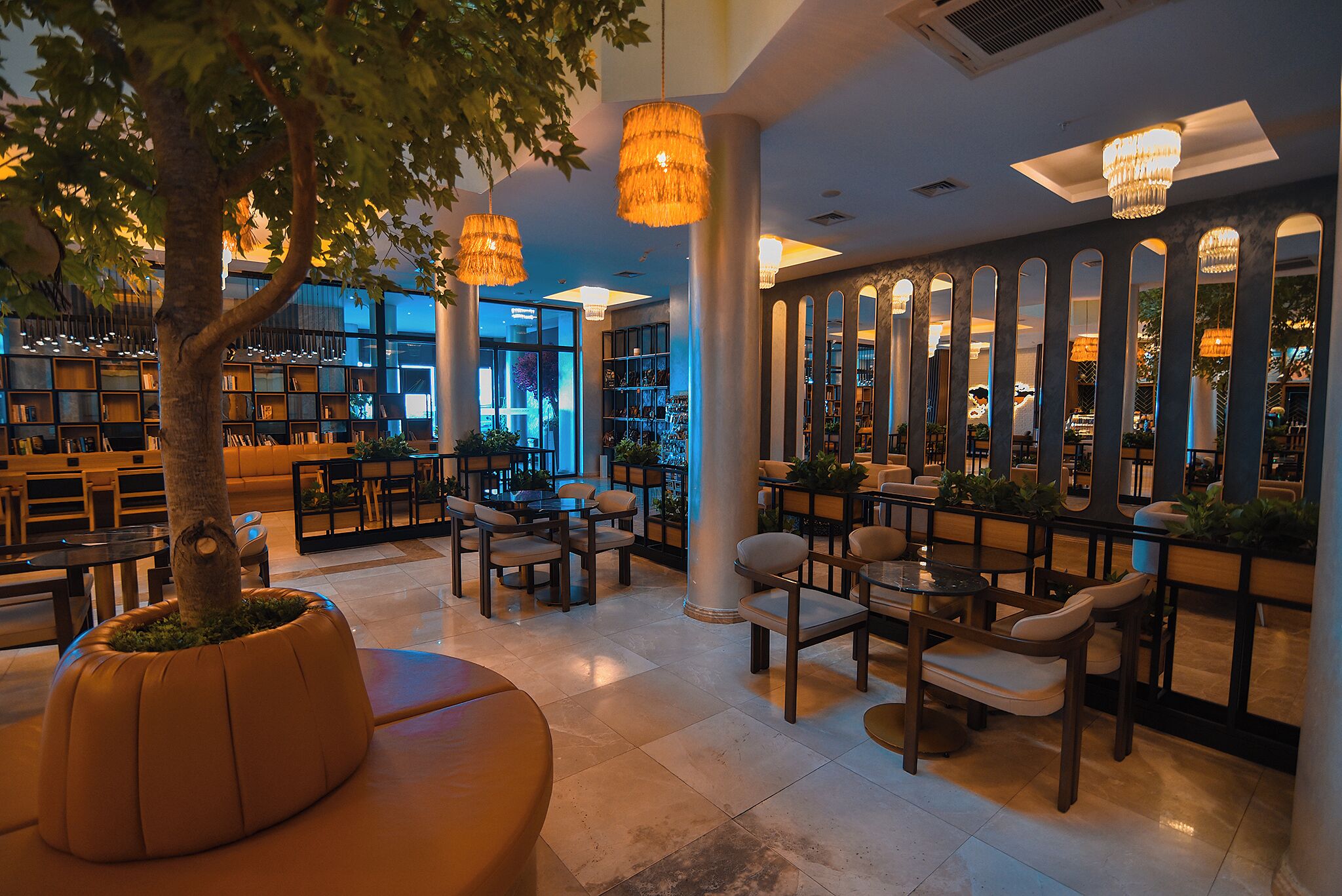 A spacious indoor area with multiple seating arrangements, including tables and chairs, surrounded by bookshelves and a large tree.