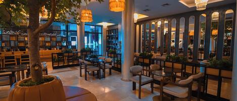A spacious indoor area with multiple seating arrangements, including tables and chairs, surrounded by bookshelves and a large tree.
